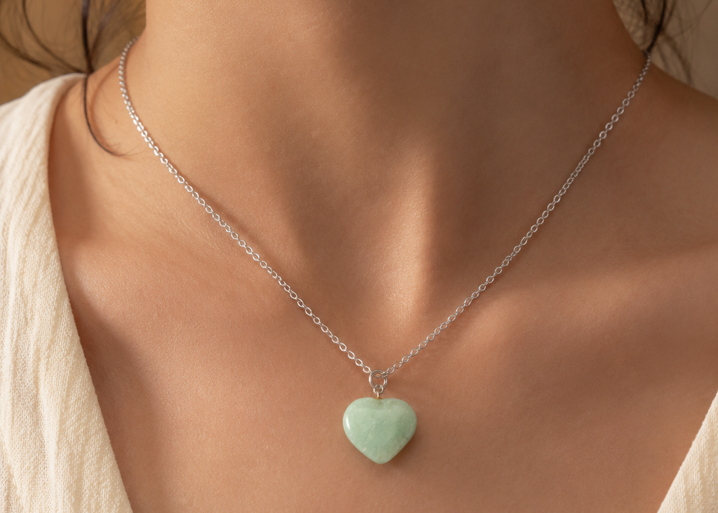 A close-up of a model's neck and chest wearing a delicate silver chain necklace with a small, smooth, light green heart-shaped pendant.